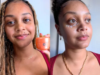 A person smiling while holding the Tatcha The Brightening Eye Cream jar near their face and under their eye 